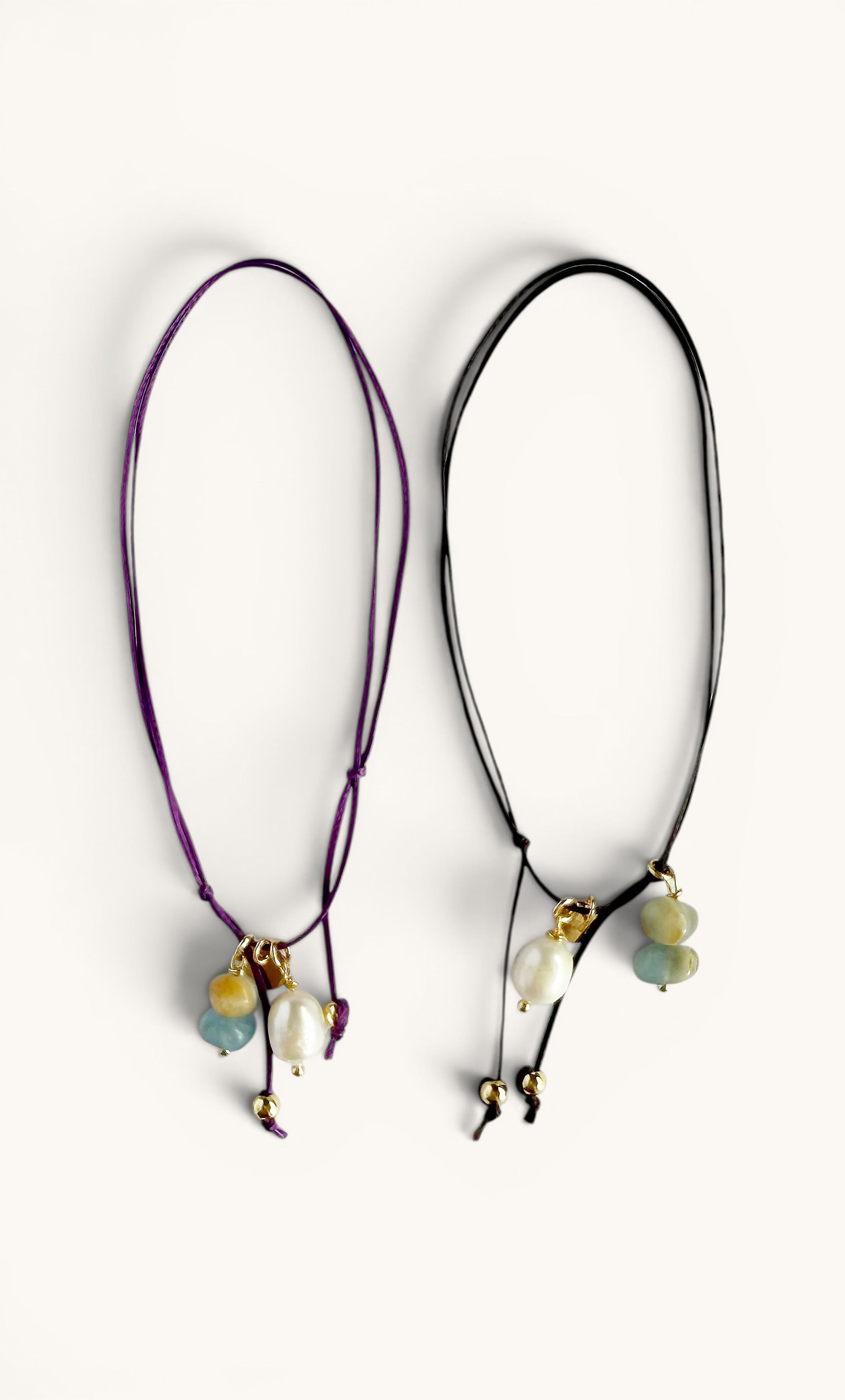 Two delicate anklets with adjustable cords, one deep purple and one black, each featuring gold-filled beads, small pearls,.