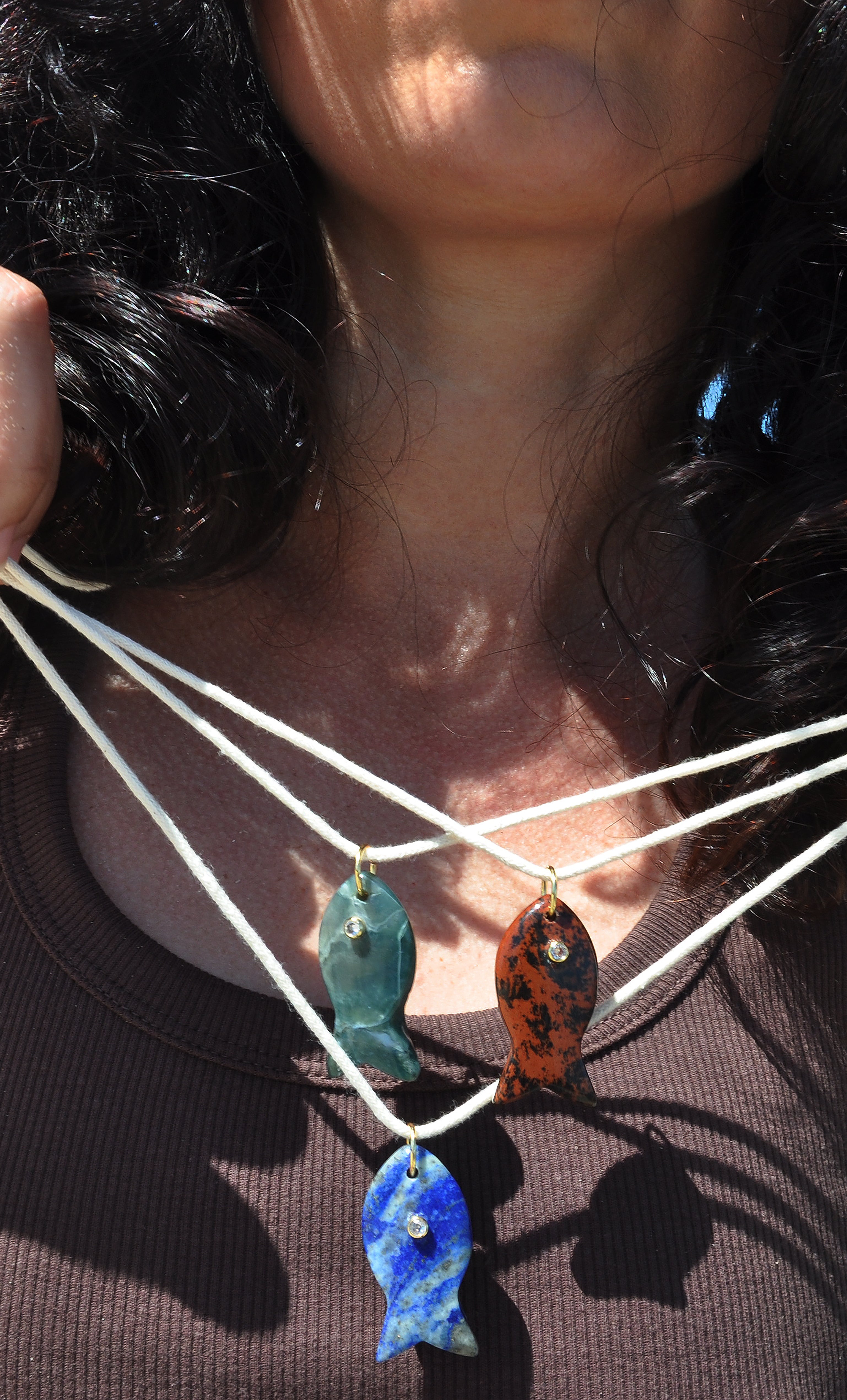 Three Tunik gold fill necklaces with carved green apatite, brown, and blue stone fish pendants on cream cords.