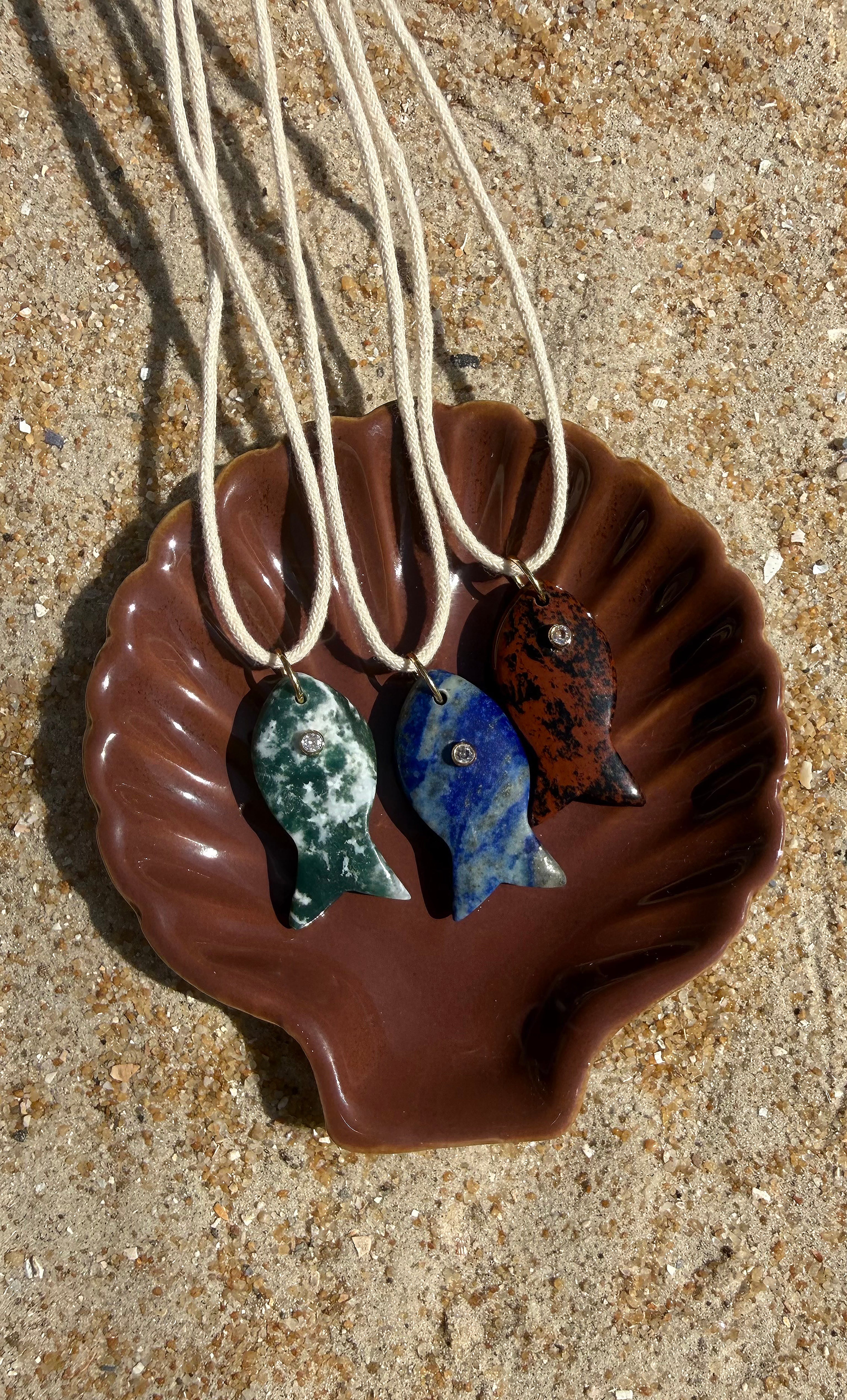 Three Tunik gold fill necklaces with carved red jasper, blue lapis, and green jasper stone fish pendants displayed on a.