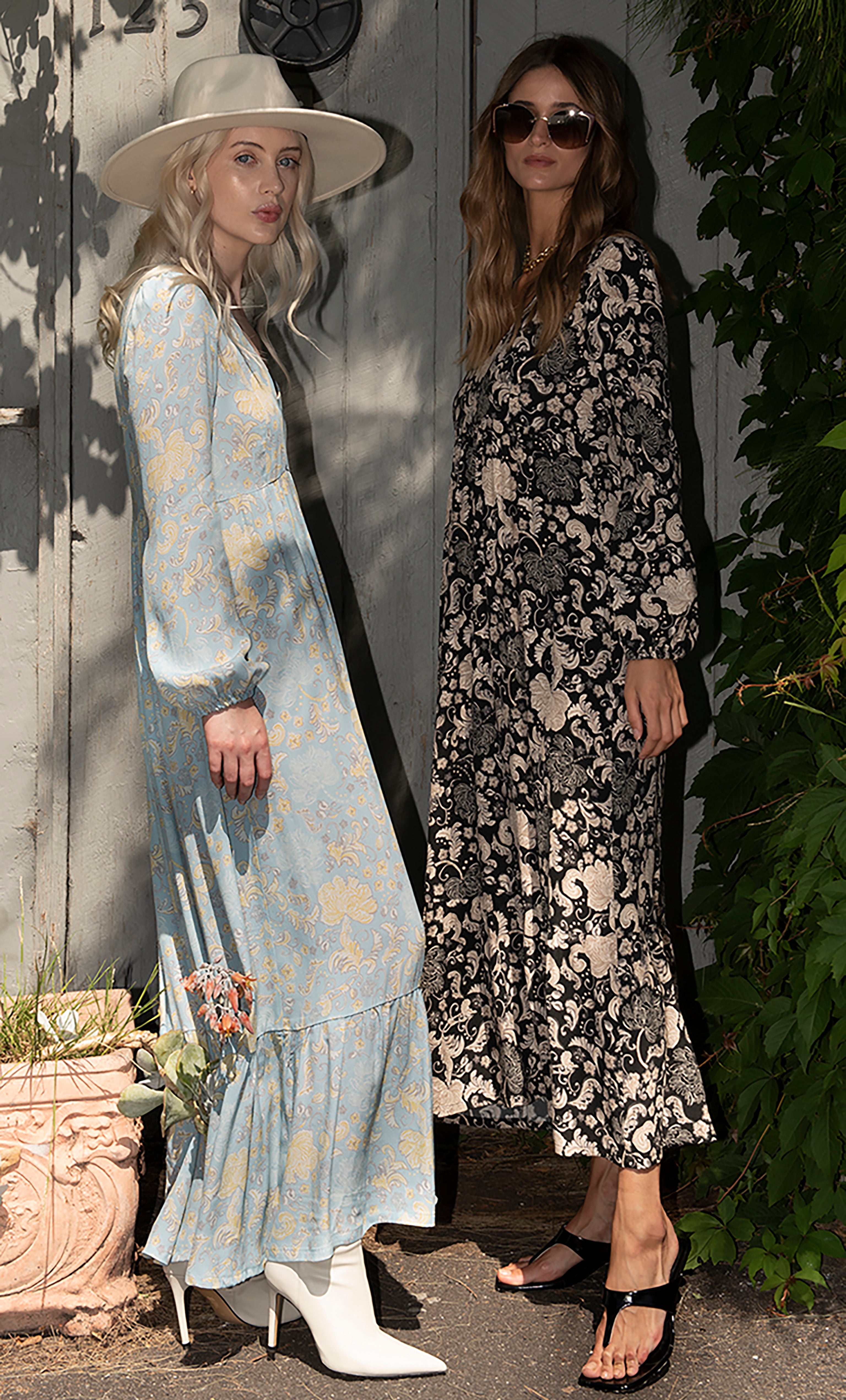 Two women wearing Tunik maxi dresses with floral prints, one in aquamarine with a gold necklace and white boots, the other.